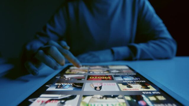 Close up of unrecognizable woman sitting at table with tablet lying down on it choosing film to watch on streaming service at night time