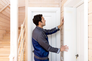 Installation of a wooden door. Portrait of young handyman in blue uniform installing door. Professional repair service and maintenance concept