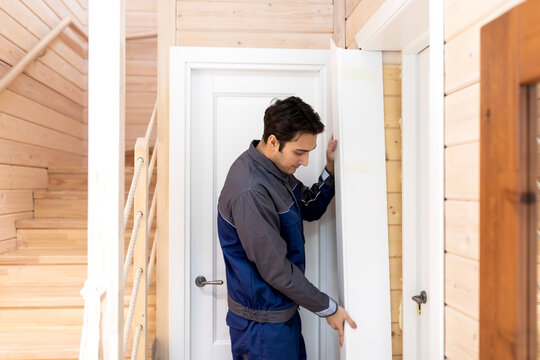 Installation of a wooden door. Portrait of young handyman in blue uniform installing door. Professional repair service and maintenance concept
