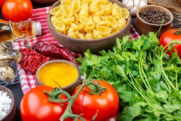 Side view of raw pastas in a brown bowl on red stripped towel yellow ginger fresh vegetables on black background