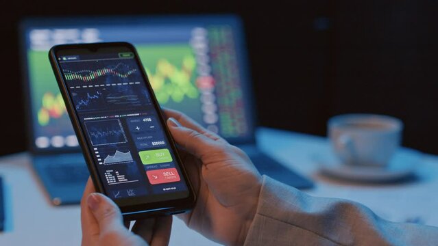 Panning left close up of unrecognizable woman sitting at desk holding smartphone with crypto app indoor at night time