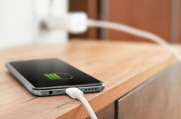 the smartphone is charged via a USB cable connected to an electrical outlet on the table - obrazy, fototapety, plakaty