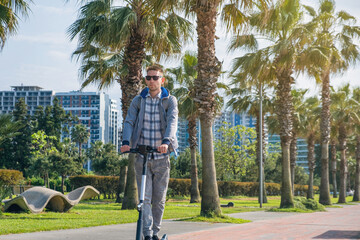Fototapeta na wymiar Young man riding electric scooter eco friendly transport along the promenade in the city with palm trees. Male driving e-scooter sustainable and outdoor in summer day. Sustainable lifestyle