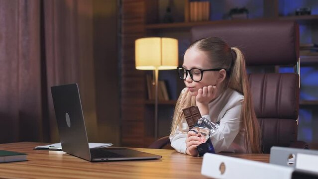 Angle view of sweet little girl relaxing in comfortable office chair and watching animated movie in luxury working place. Adorable schoolchild relishing bar of milk chocolate and waiting for parents.