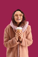 Stylish young woman in teddy coat with cup of coffee on pink background