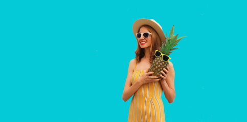Summer portrait of happy smiling woman with pineapple wearing sunglasses, straw hat posing on blue background