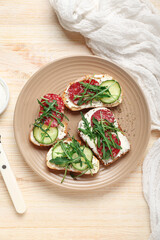 Plate of tasty sandwiches with cream cheese, cucumber, arugula and salami on light wooden table