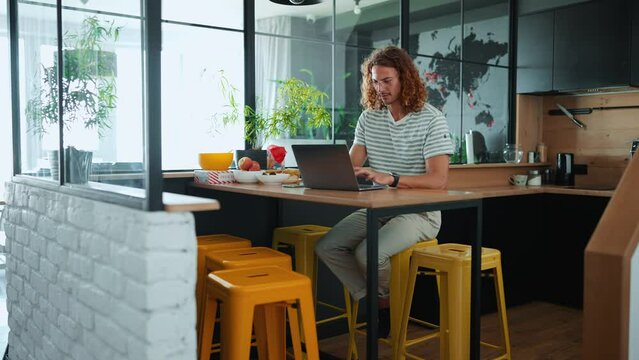 Smiling curly haired man in eyeglasses working on laptop in office kitchen 