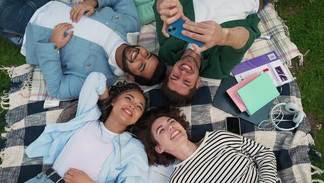 Top view of positive multiethnic friends take selfies on mobile while lying on blanket at picnic in park