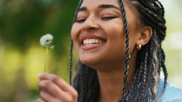 Smiling african woman with braided pigtails blows on dandelion and makes wish in park