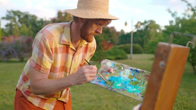 The artist paints a picture in the park. Green landscape, grass trees and flowers. The easel on which the painting stands. Blue, green colors in the palette.