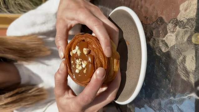 Close-up of female hands tearing a delicious cruffin with salted caramel into halves, caramel dripping down the fingers, female sitting outside at a glass table of a cafe terrace. Lifestyle vertical v