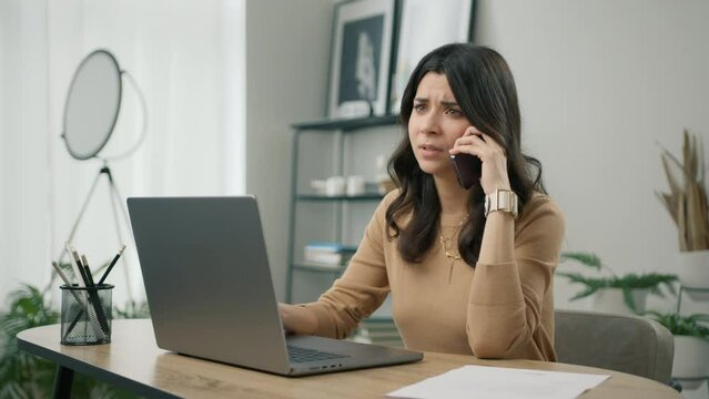 Portrait of displeased young hispanic woman looking at laptop screen, dissatisfied with bad news message during smartphone call. Unhappy stressed female mobile user hearing bad news at mobile chat 4K