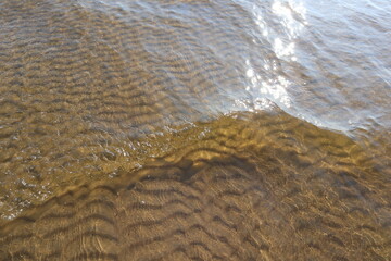 Transparent wave on sea water with sun glare and ripples on sandy bottom. Sea waves and gritty surface texture created a pattern of dunes on sand of bottom.