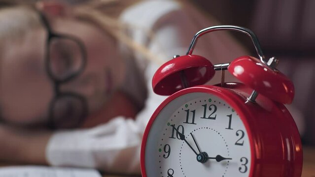 Close up of red manual alarm clock set for 3 PM in office place and peaceful little girl sleeping at table on blurred background. Cute female kid in black glasses having short break and taking nap.