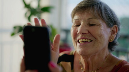 Happy older woman waving hello or goodbye while holding cellphone device at home speaking by video...