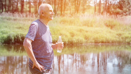 Active older male person standing on riverbank with water bottle. Senior human lifestyle and leisure concept.
