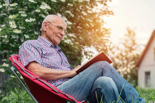 Retired man seated on armchair wearing glasses reading book in garden outdoor of cottage house.
