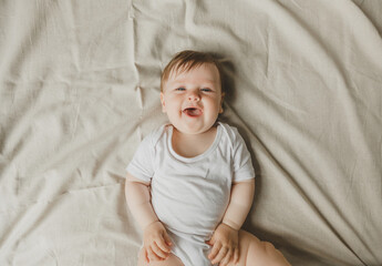 Charming blue-eyed 6-month-old baby lies in bed in a white bodysuit. View from above