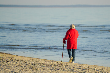 An elderly woman in a red jacket with walking stick walks along the seashore