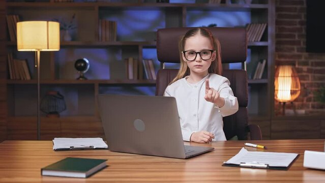 Front view of severe little girl sitting in front of personal laptop and waving finger in disagreement. Displeased kid in white shirt and glasses showing dissatisfaction with opponent words in office.