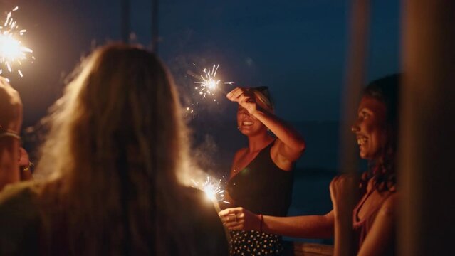 Diverse women dance and laugh with sparklers at seaside tropical hen party at sunset. Young smiling multiracial females have fun at Independence Day celebration by the sea with sparkling lights