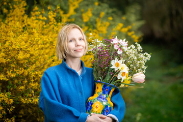 Attractive adult woman in autumn or spring in a blue coat with a bouquet of various flowers against the backdrop of a blooming yellow shrub.