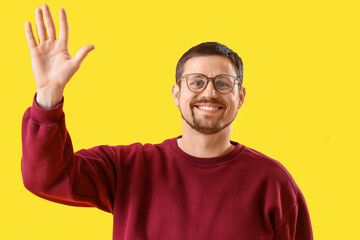 Handsome man in stylish eyeglasses waving hand on yellow background