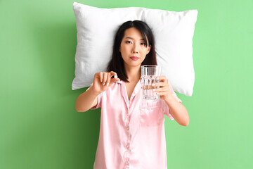 Beautiful Asian woman in pajamas with glass of water and pill on green background