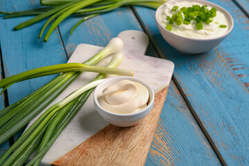 Fototapeta na wymiar Board with bowls of tasty sour cream and green onion on blue wooden background