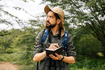 Man in nature with hat, backpack, and binoculars taking field notes - obrazy, fototapety, plakaty