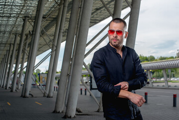 a stylish man in light pants and a black shirt against the background of empty walls holds glasses in his hands and looks away angrily