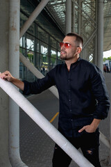 a stylish man in light pants and a black shirt against the background of empty walls holds glasses in his hands and looks away angrily