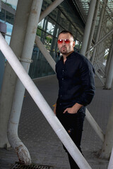 a stylish man in light pants and a black shirt against the background of empty walls holds glasses in his hands and looks away angrily