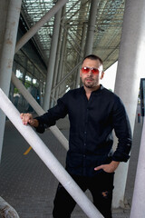 a stylish man in light pants and a black shirt against the background of empty walls holds glasses in his hands and looks away angrily