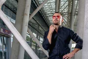 a stylish man in light pants and a black shirt against the background of empty walls holds glasses in his hands and looks away angrily