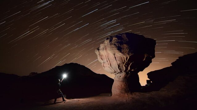 man standing near a rock mushroom and lighting during the night with startrails in the back ground 