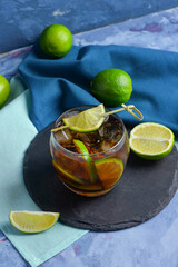Board with glass of cold Cuba Libre cocktail and limes on blue background