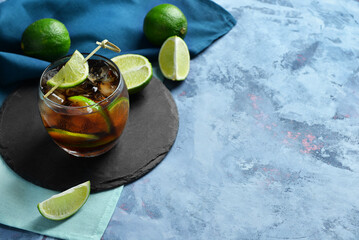 Board with glass of cold Cuba Libre cocktail and limes on blue background