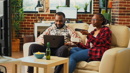 People watching movie on tv together in living room, browsing internet on laptop and eating asian food with chopsticks. Modern couple feeling relaxed on couch with takeout meal. Tripod shot.