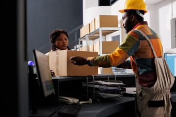 Postal service worker managing parcel receiving and registration in warehouse. African american woman courier putting cardboard box on counter desk in shipment company storehouse