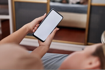 Closeup of young woman holding smartphone with white screen mockup while lying in bed and scrolling social media