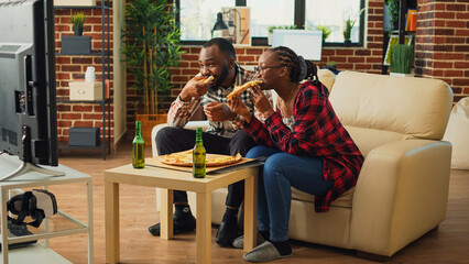 Fototapeta na wymiar Happy partners eating slices of pizza on sofa and binge watching tv show at home, enjoying fast food meal. Modern couple receiving takeaway food order and sitting to have dinner at television.