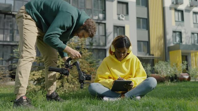 Professional videographer with camera in his hands shoots dark-skinned girl sitting on lawn with headphones and tablet in her hands on territory of residential complex. Backstage. Slow motion