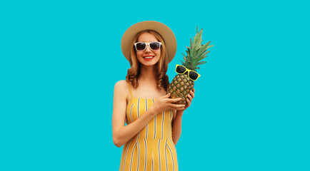Summer portrait of happy smiling woman with pineapple wearing sunglasses, straw hat posing on blue...