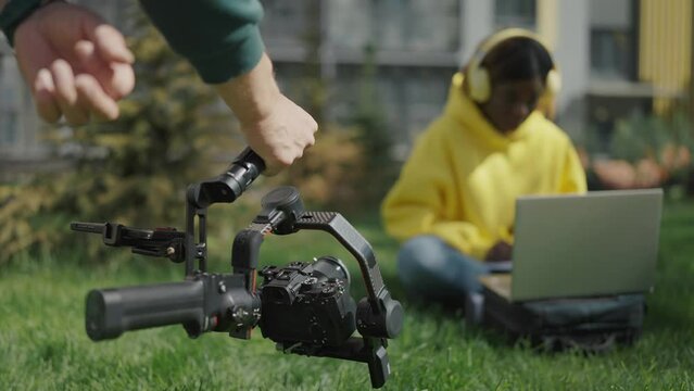  Close-up of operator hands holding camera and filming african girl who is sitting at laptop on lawn in courtyard of residential complex. Backstage. Slow motion
