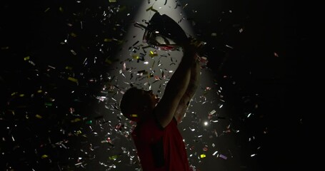 Silhouette of Caucasian male rugby player raising a trophy above head against bright light and falling confetti. Super slow motion, shot on RED cinema camera - Powered by Adobe