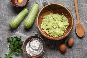 Bowl with grated zucchini and ingredients for preparing fritters on grey background