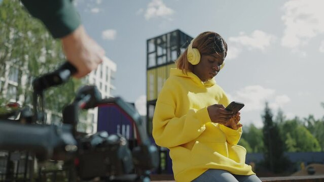 Videographer shoots african american woman in yellow hoodie wearing headphones with smartphone in her hands on bench of residential complex. Backstage. Slow motion