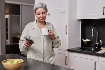 Waist up portrait of modern mature woman using smartphone at kitchen in morning and drinking coffee, copy space